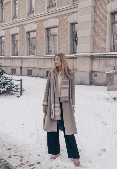 Mujer con abrigo beige, bufanda, guantes y pantalones negros parada en una acera nevada junto a un edificio de ladrillo con varias ventanas.