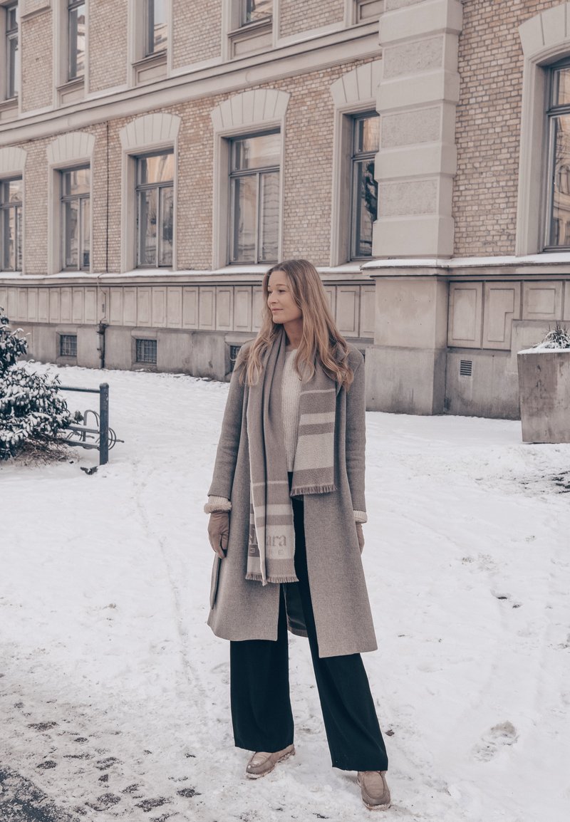 Kvinde i beige frakke, tørklæde, handsker og sorte bukser står på en sneklædt fortov ved siden af en murstensbygning med flere vinduer.