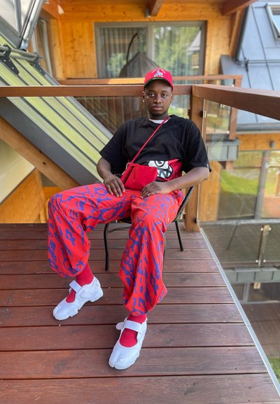 Pantalones cargo rojos con un patrón, camiseta negra, bolsa de mano roja, zapatos deportivos blancos con correa y calcetines rojos. Sentado en una silla de patio de madera.