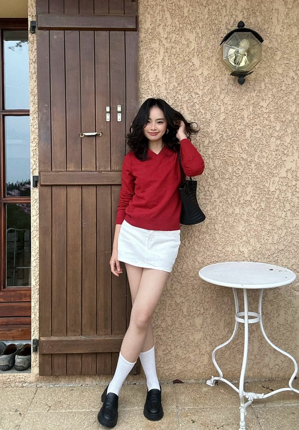 Une femme vêtue d’un pull rouge, d’une jupe blanche et de chaussures noires pose à côté d’une porte en bois et d’une petite table de patio blanche.