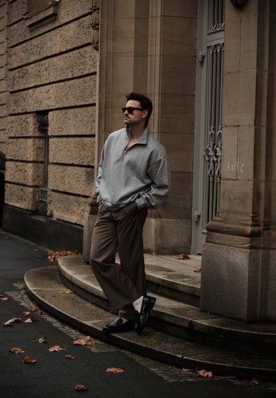 Sudadera gris con cremallera en el cuello, pantalones marrones plisados, mocasines negros y gafas de sol, posando contra una pared de piedra con hojas otoñales en el pavimento.