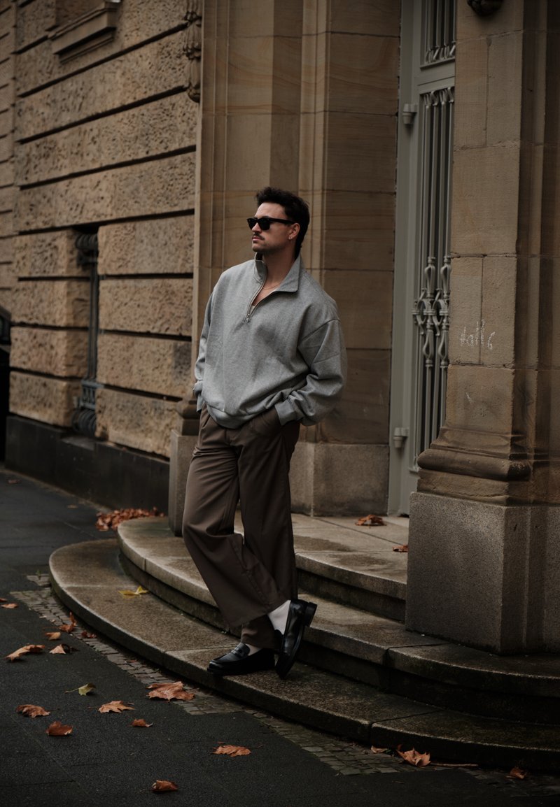 Sivi duks s patentnim zatvaračem na vratu, smeđe plisirane hlače, crne mokasinke i sunčane naočale, pozirajući uz kameni zid s jesenjim lišćem na pločniku.