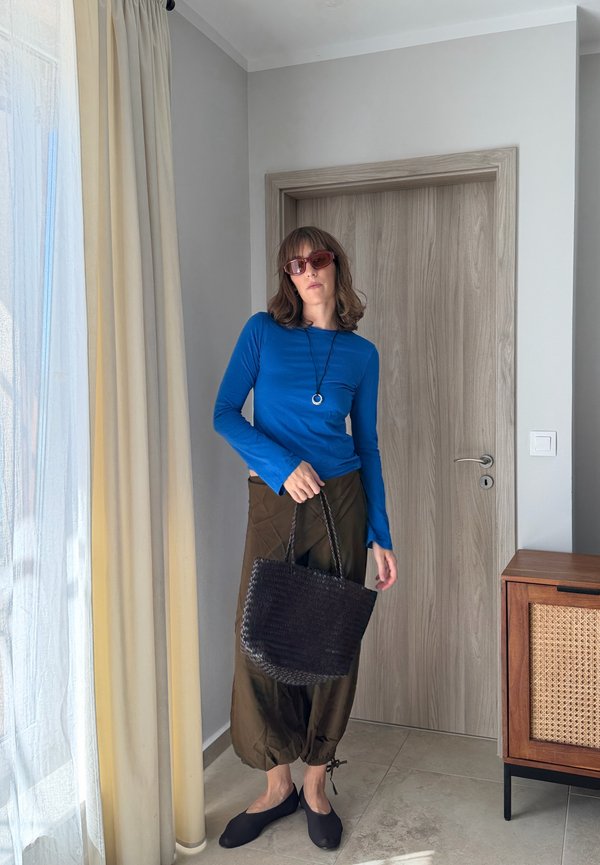 Mujer con camiseta azul de manga larga, pantalones marrones sueltos, zapatos planos negros, gafas de sol y collar, sosteniendo un bolso negro tejido en una habitación con puerta de madera y cortina.