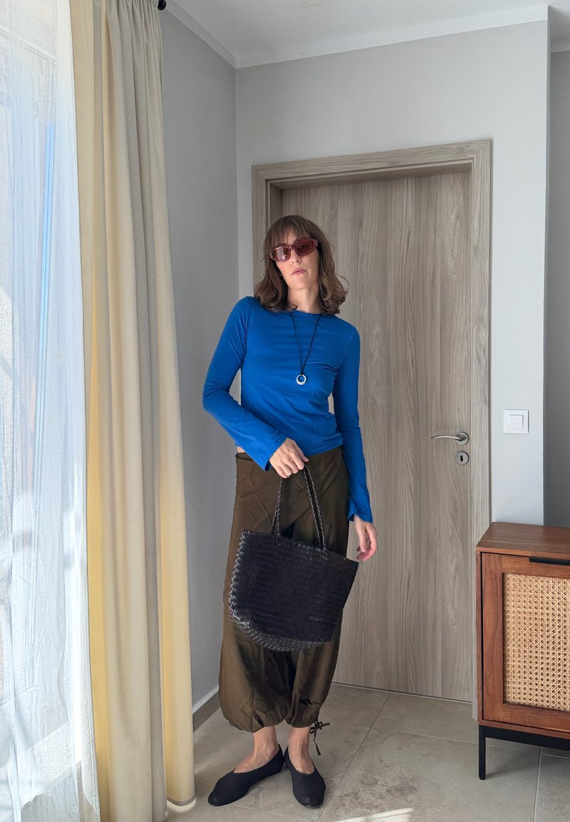 Femme portant un haut bleu à manches longues, un pantalon marron ample, des ballerines noires, des lunettes de soleil et un collier, tenant un sac noir tressé dans une pièce avec une porte en bois et un rideau.