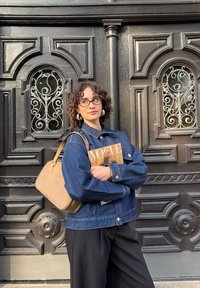 Denim jaka ar zilām nokrāsām, ar pogām slēdzama, lielā izmēra, apvienota ar melniem biksēm un ķieģeļu krāsas rokassomu, turot žurnālu "VOGUE".