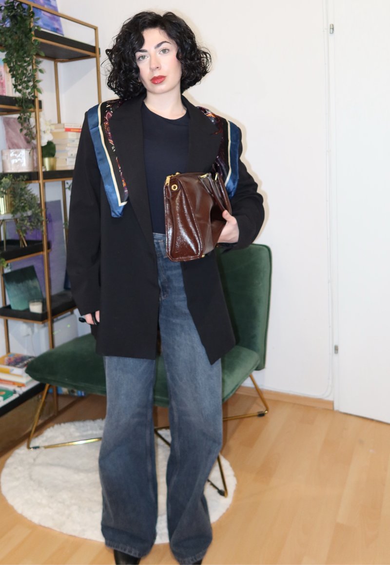 Blazer preto oversize com lenço estampado, camisa escura, jeans de ganga de perna larga, segurando uma carteira castanha, em pé numa cadeira verde.