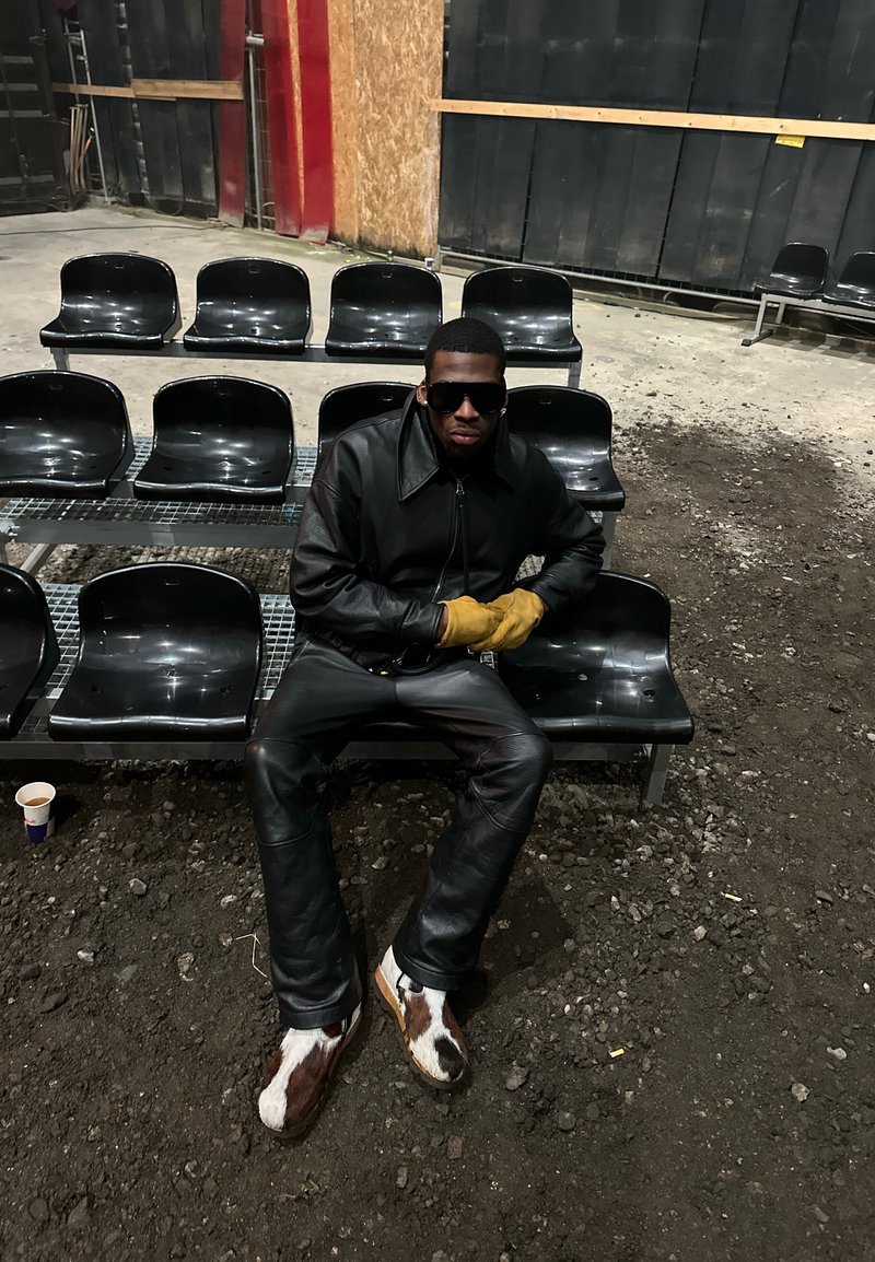 Homme portant une veste en cuir noire, un pantalon, des lunettes de soleil et des gants jaunes, assis sur des chaises en plastique noires dans un espace intérieur faiblement éclairé, recouvert de gravier.