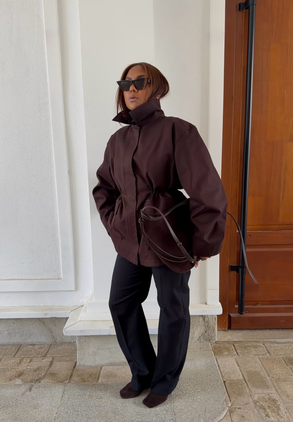 Una mujer con gafas de sol posa junto a una entrada. Lleva una chaqueta oscura y holgada, pantalones y un bolso al hombro.
