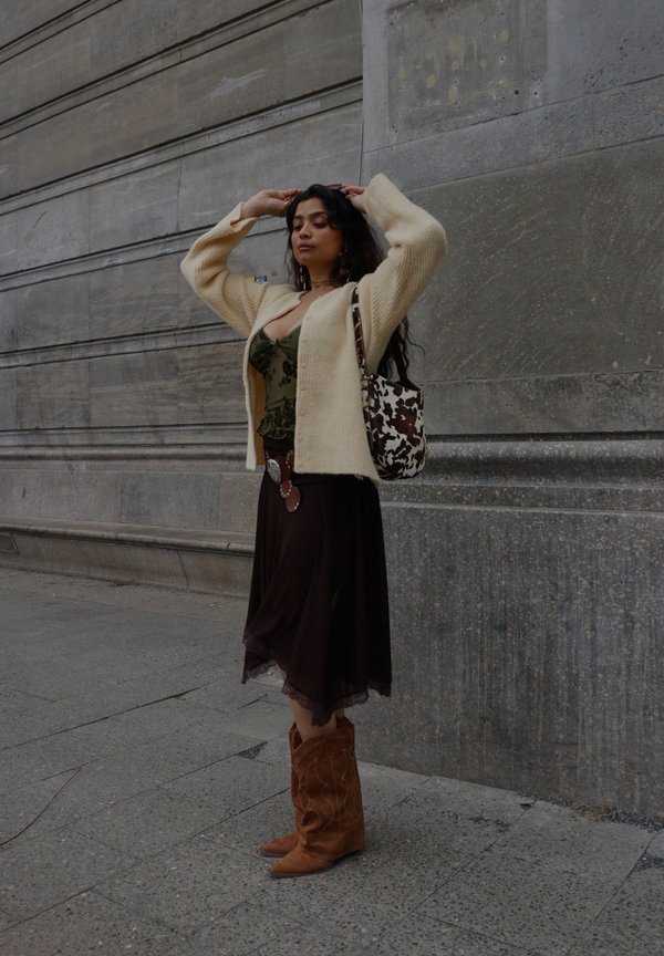 Žena s dlhými tmavými vlasmi si upravuje sveter, má na sebe zelený top, hnedú sukňu, svetlohnedé čižmy a nesie tašku s motívom kravy, stojac pred kamennou stenou.