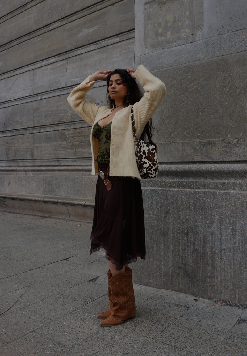 Γυναίκα με μακριά σκουρόχρωμα μαλλιά ρυθμίζει το ζακέτα της, φοράει πράσινο τοπ, καφέ φούστα, ταμπά μπότες και κρατά τσάντα με μοτίβο αγελάδας μπροστά σε πέτρινο τοίχο.