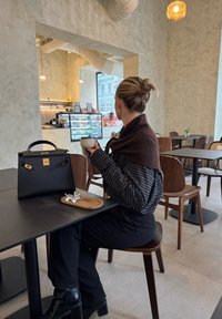 Femme avec un chignon tenant une tasse, assise à une table de café avec un sac à main noir et des écouteurs sur un plateau dans un intérieur minimaliste et moderne.