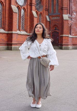 Blusa blanca de ojo de perdiz con volantes, falda gris fluida y bolso de mano beige. De pie sobre un pavimento gris frente a un edificio de ladrillo.