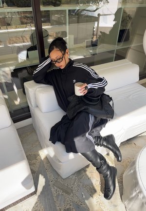 Mujer con chándal negro de Adidas y botas sentada en un sofá blanco, sosteniendo una taza de café, con un bolso negro sobre sus piernas, en el interior.