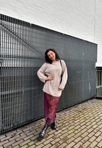 Suéter bege canelado de mangas longas, combinado com uma saia burgundy transparente e botas pretas robustas, contra um fundo cinzento com tábuas verticais.