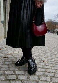 Μαύροι πλατφόρμες loafers με ασημένια τρουκς, φοριούνται με μαύρες καλσόν και πλισέ μαύρη φούστα. Ένα μικρό κόκκινο τσαντάκι κρατιέται με το ένα χέρι.