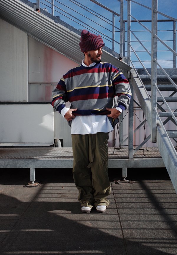 Gestreepte sweatshirt in grijs, rood en geel, laag over een wit shirt, gecombineerd met losse olijfgroene broeken en witte schoenen.