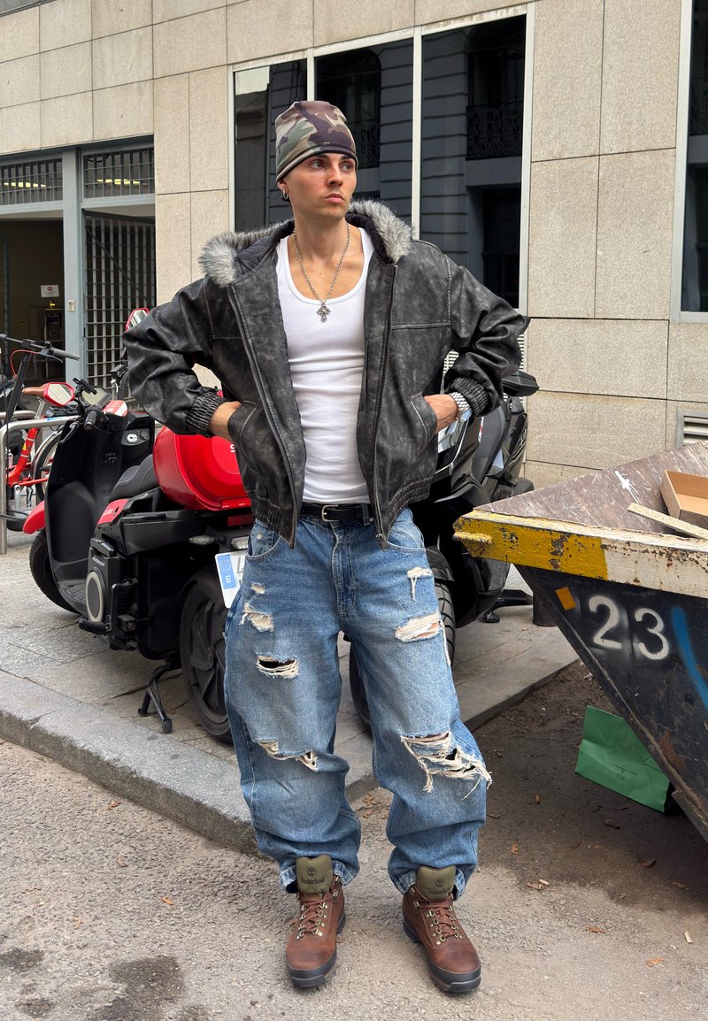 Læderbomberjakke med pelsforet hætte, hvid tanktop, slidte blå jeans og brune støvler. Urban baggrund med parkerede scootere.