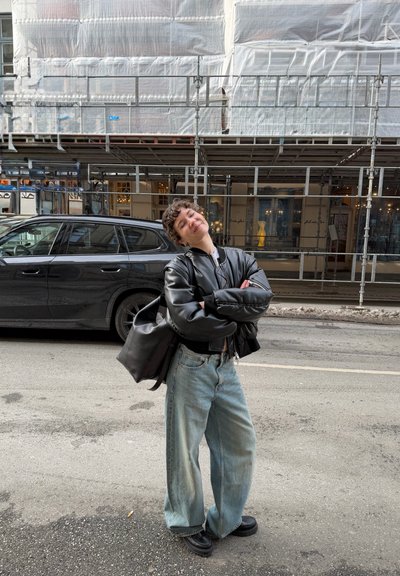 Persona con cabello corto y rizado, con chaqueta de cuero negra y pantalones vaqueros anchos, de pie sonriendo en una calle de la ciudad frente a un edificio enandamado y un coche aparcado.
