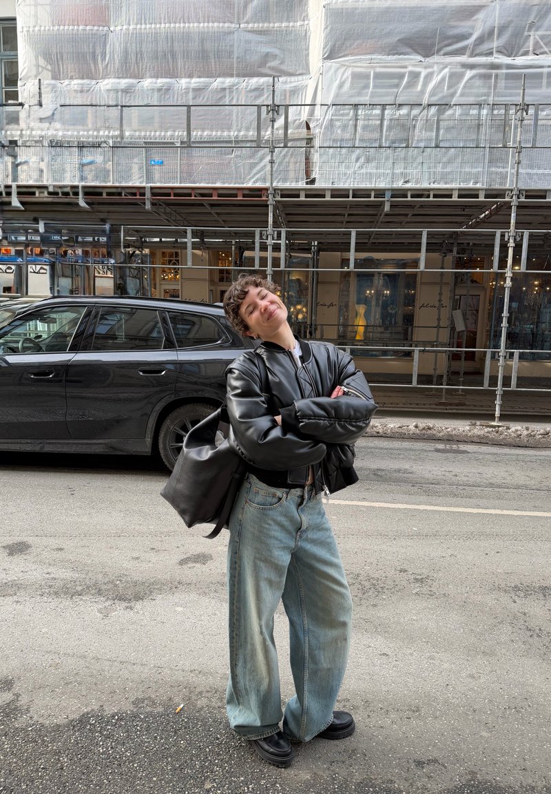 Personne aux cheveux courts et bouclés, vêtue d'une veste en cuir noire et d'un jean large, souriant dans une rue de la ville devant un bâtiment échafaudé et une voiture garée.