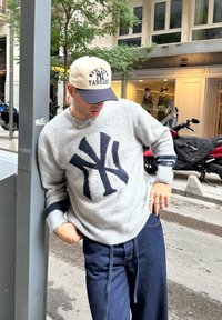 Grijze gebreide trui met een donkerblauw New York Yankees-logo, gecombineerd met donkere denim broek. De outfit bevat een beige en marineblauwe pet en zilveren sieraden.