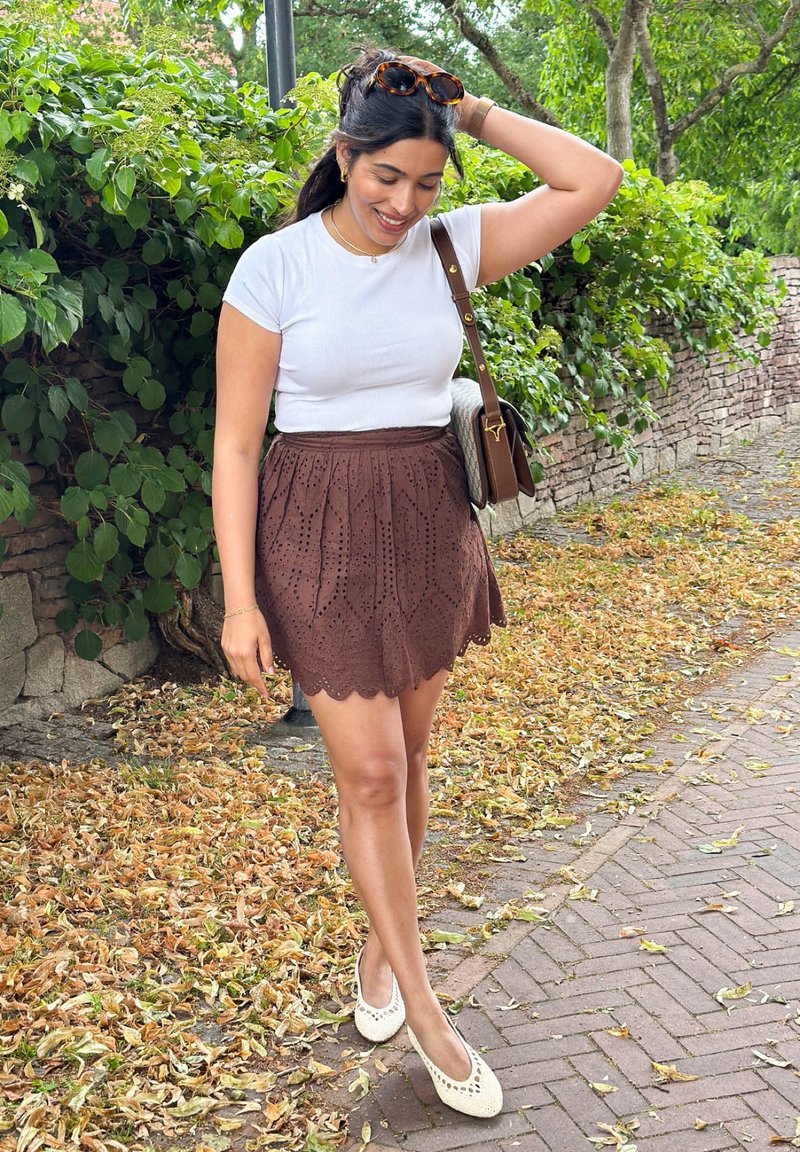 Young woman in white top and brown skirt stands on leaf-covered path, smiling and touching hair in green outdoor setting.