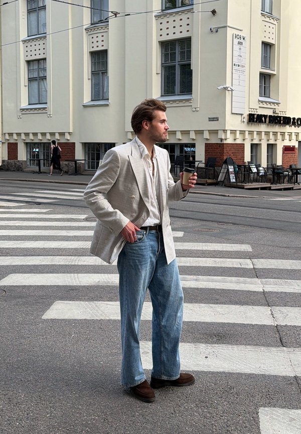 Homme en blazer beige et jean tenant un café, debout sur un passage pour piétons devant un café dans une rue urbaine.