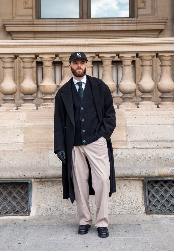 Partasuinen mies, jolla on musta ylipuku, musta neuletakki, valkoinen paita, musta solmio, beiget housut, mustat käsineet ja musta NY-lippalakki, seisoo kiven kaiteen vieressä.