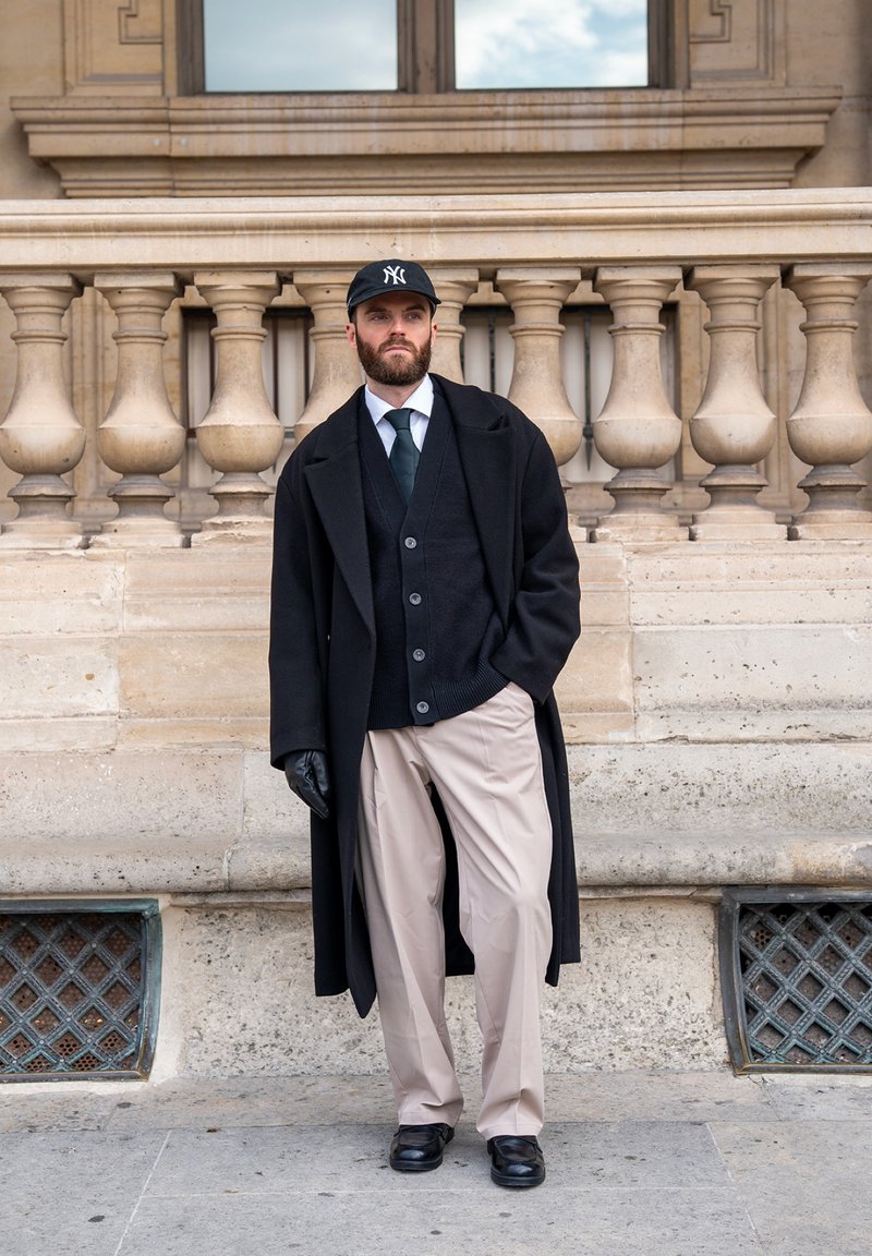 Homme barbu portant un manteau noir, un cardigan noir, une chemise blanche, une cravate noire, un pantalon beige, des gants noirs et une casquette de baseball noire NY, debout près d'une balustrade en pierre.
