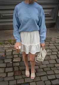 Felpa azzurra chiara con testo ricamato, sovrapposta a una gonna bianca in pizzo con volants. Mule beige e borsa chiara. Tazza di caffè in mano.