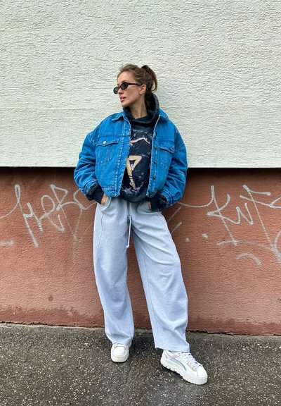 Chaqueta de denim azul, sudadera gráfica negra debajo, pantalones de chándal grises y zapatillas blancas de plataforma. El fondo presenta graffiti en una pared.
