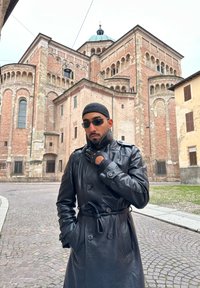 Homme portant un manteau en cuir noir, des gants sans doigts et des lunettes de soleil, debout devant un grand bâtiment historique en briques avec des fenêtres en arc.