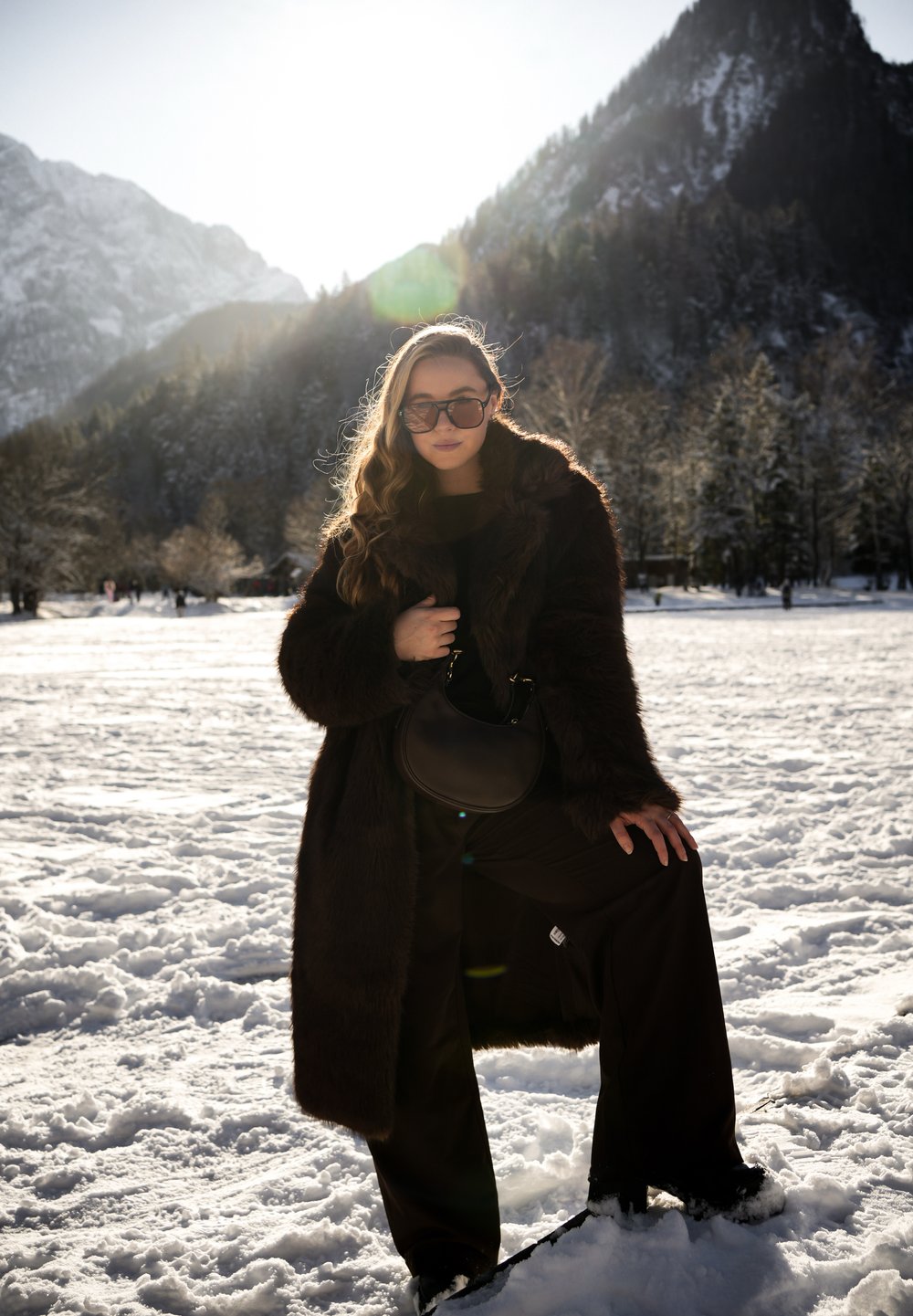 Γυναίκα που φοράει σκουρόχρωμο γούνινο παλτό, γυαλιά ηλίου και μαύρο παντελόνι, στέκεται στο χιόνι με το ένα πόδι σηκωμένο, με βουνά και δέντρα στο φόντο.