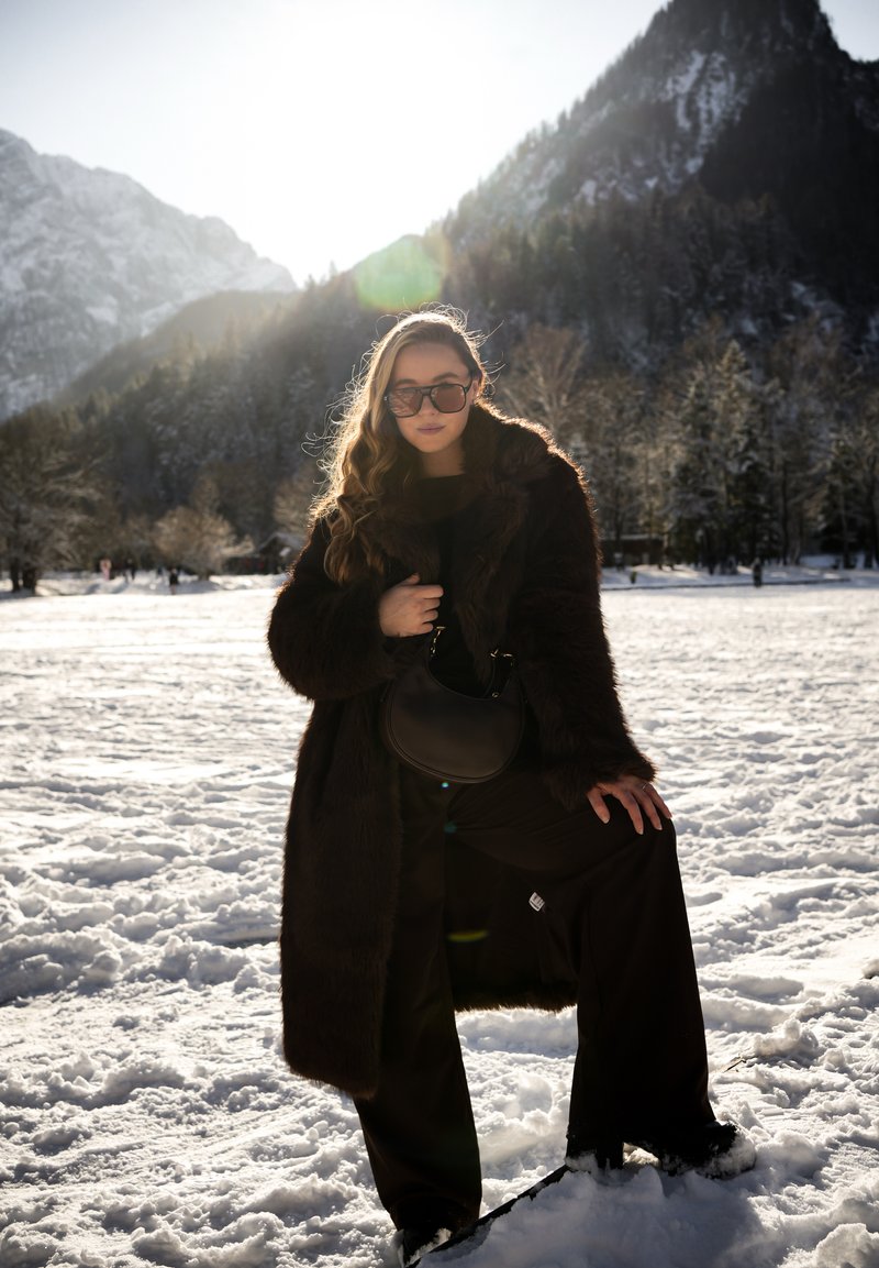 Femeie purtând o haină de blană închisă la culoare, ochelari de soare și pantaloni negri stă pe zăpadă cu un picior ridicat, munți și copaci în fundal.