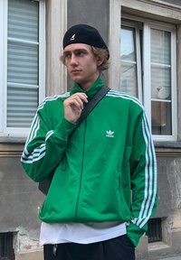 Zielona kurtka adidas z białymi paskami, z zamkiem z przodu, noszona na białej koszuli. Widoczna jest także czarna czapka i torba na ramię.