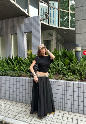 Top corto negro con falda larga negra de cintura alta y cinturón beige. Los accesorios incluyen un brazalete plateado y un anillo redondo. El fondo presenta vegetación y azulejos.