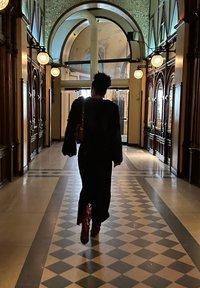 Μαύρο μακρύ φόρεμα με υφασμάτινα μανίκια, καφέ μπότες και τσάντα ώμου, περπατώντας σε ένα μοτίβο με μαύρα και άσπρα πλακάκια σε έναν διάδρομο.