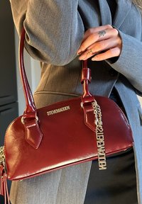 Burgunderfarbene Lederhandtasche mit kurzem Henkel, goldfarbenen Beschlägen und einer dekorativen Kette. Mit einem auf der Vorderseite geprägten Logo. Glatte Textur.