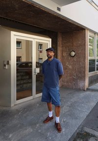 Mand iført blå skjorte, denimshorts, kasket og briller står på fortovet foran en bygningse entrance med glasdøre og sten vægge.