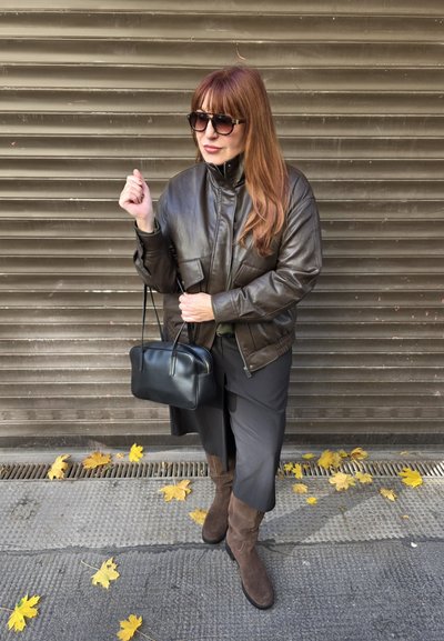 Chaqueta bomber de cuero marrón, bolso negro, botas de ante marrón hasta la rodilla, con gafas de sol, falda oscura y suelo texturizado con hojas de otoño.