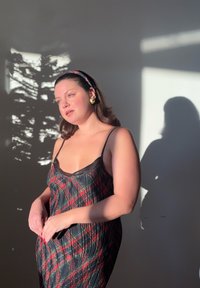 Vestito a trapezio a quadri neri con strisce rosse e verdi, rifinitura nera trasparente e orecchini dorati. L'illuminazione soffusa crea ombre sulla parete.