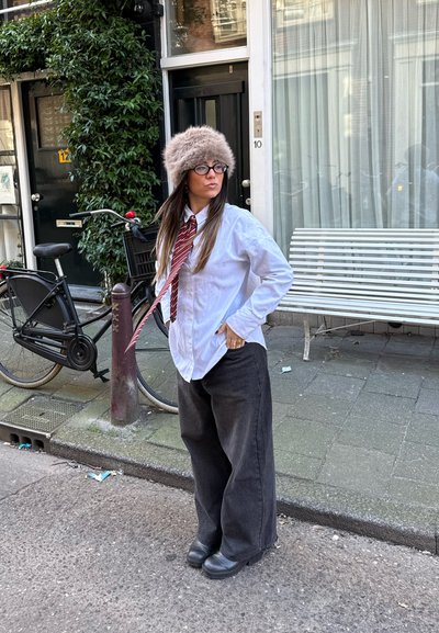 Camisa blanca de botones, corbata a rayas burdeos, pantalones anchos negros, sombrero de piel gris; de pie en una calle con una bicicleta negra cerca.