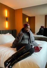 Femme aux cheveux roux en veste grise et bottes noires assise sur un lit blanc dans une chambre d'hôtel moderne avec des murs en bois et des lumières verticales.
