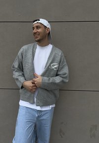 Pelēks adīts kardigans ar baltu izšuvuma logo, valkāts virs baltas T-krekla. Gaismas zili džinsi pabeidz ikdienas tēlu. Fons ir neitrāls.