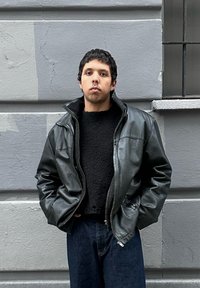 Νεαρός με κοντά σκούρα μαλλιά που φοράει μαύρο δερμάτινο μπουφάν και μαύρο πουλόβερ στέκεται μπροστά σε γκρίζο τσιμεντένιο τοίχο με παράθυρο.