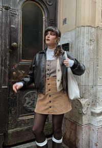 Brun læder bomberjakke over en houndstooth-mønstrede top og en sandfarvet knap-front nederdel. Hvid taske og knæhøje støvler med hvid trim.