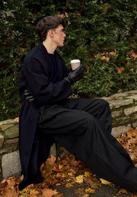 Soprabito nero e pantaloni larghi neri, abbinati a guanti neri. Tiene in mano una tazza di caffè, seduto su un muro di pietra circondato da foglie autunnali.