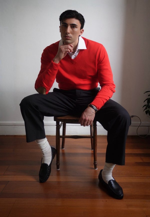 Man in rode trui en wit overhemd zit op een houten stoel met gespreide benen, draagt zwarte broek, witte sokken en zwarte loafers op een houten vloer.