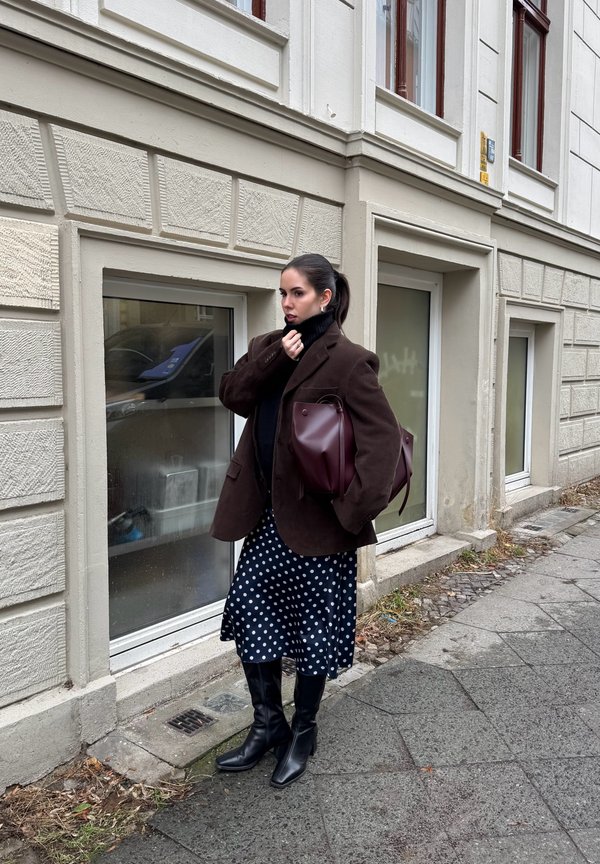 Γυναίκα που στέκεται σε πεζοδρόμιο δίπλα σε μια μπεζ οικοδομή, ντυμένη με καφέ σακάκι, μαύρο ζιβάγκο, φούστα με πουά, μαύρες μπότες και κρατώντας μια μεγάλη τσάντα.