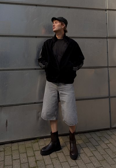 Chaqueta negra con cremallera y rayas, camiseta negra, pantalones cortos de mezclilla grises, botas de tobillo marrones, con un fondo metálico y pavimento de adoquines.