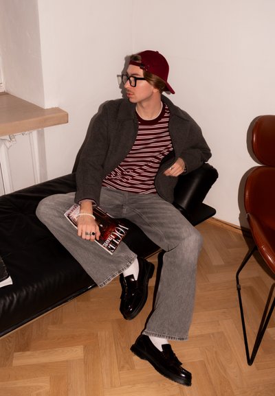 Joven con gafas, gorra burdeos, camiseta a rayas y chaqueta gris sentado en un sofá negro sosteniendo una revista en una habitación minimalista.